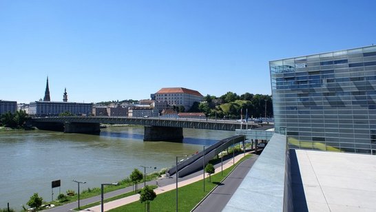 Nibelungenbruecke Linz Ein Fluss mit einer Brücke darüber. Am Fluss entlang gibt es einen Fußgänger und Radweg. Rechts ist das Ars Electronica Center.